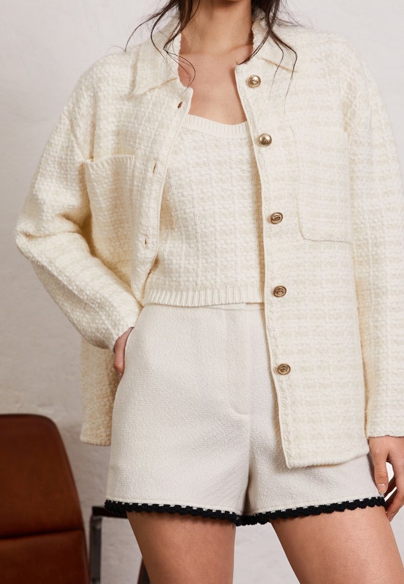 Femme portant une veste boutonnée texturée couleur crème, un crop top en maille assorti et un short crème avec une bordure noire, debout à l'intérieur.