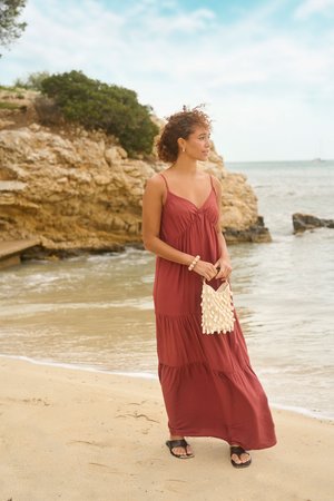 Frau trägt ein rostfarbenes Maxikleid und Sandalen, steht an einem sandigen Strand und hält eine mit Muscheln verzierte Handtasche nahe felsigen Klippen.