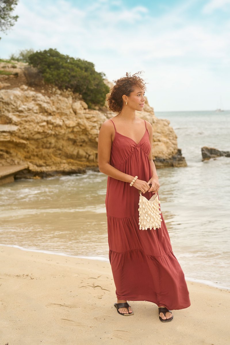 Femme portant une robe longue couleur rouille et des sandales, debout sur une plage de sable tenant un sac décoré de coquillages près de falaises rocheuses.