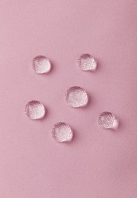 Sześć przezroczystych kropli wody spoczywających na teksturowanej różowej tkaninie, pokazujących odporność na wodę z brakiem wchłonięcia lub rozprzestrzenienia.