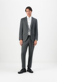 Młody mężczyzna stoi na białym tle, ubrany w szary garnitur z dopasowanymi spodniami, białą koszulę z guzikami oraz czarne eleganckie buty.
