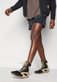 Αθλητικά παπούτσια με ψηλό ποδόγυρο σε μαύρο και μπεζ σχέδιο, με δίχτυ ψηλά και υφή στην σόλα, συνδυασμένα με σορτς με σχέδιο και καφέ μπλούζα.