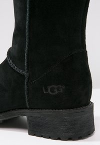 Bottine UGG en daim noir avec coutures visibles et semelle robuste, gros plan montrant le talon et le logo.