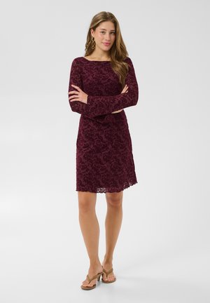 Une femme présente une robe longue à manches longues, à motif bordeaux, avec des boucles d’oreilles dorées et des sandales beiges. Elle se tient les bras croisés devant un fond de studio uni.