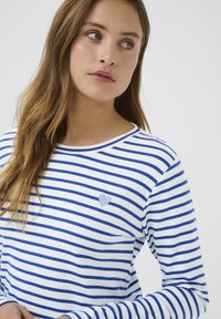 Chemise à rayures à manches longues blanche et bleue avec un col rond et une petite broderie en forme de cœur bleu sur la poitrine gauche. Tissu doux au toucher.