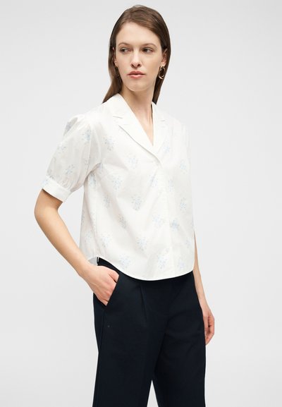 Γυναίκα που φοράει λευκό μπλουζάκι με κοντά μανίκια και φωτεινό μπλε λουλουδάτο μοτίβο και μαύρο παντελόνι, κοιτάζοντας προς τα αριστερά της με το δεξί χέρι στην τσέπη.