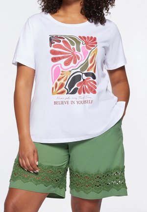 Valkoinen puuvillainen t-paita, jossa on värikäs kukkakuosi ja teksti "USKO ITSEESI"; yhdistetty vihreisiin shortseihin, joissa on pitsireunus.