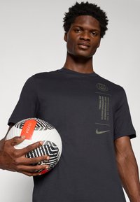 Černé tričko s krátkým rukávem a zeleným logem Nike a textem. Osoba drží vzorovaný černobílý fotbal s červenými a oranžovými akcenty.