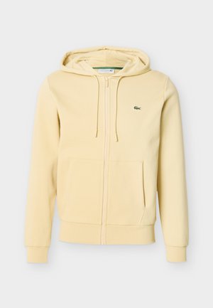 Felpa con cappuccio beige chiaro con zip, tasche frontali, cappuccio con coulisse, polsini a coste e piccolo logo del coccodrillo Lacoste verde sul petto.
