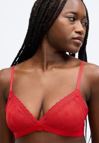 Bralette vermelho com renda, design em malha transparente, alças ajustáveis e faixa inferior lisa com detalhe de logótipo.
