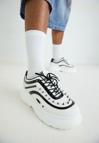 Zapatillas gruesas en blanco y negro con acentos de picos, lados texturizados y cordones negros. Llevadas con calcetas blancas hasta la rodilla.