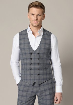 Mężczyzna ubrany w szarą kratowaną dwurzędową kamizelkę i pasujące spodnie na białej koszuli, stojący na neutralnym tle.