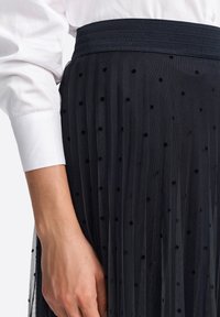 Schwarzer, plissierter Rock aus strukturiertem Stoff mit Polka-Dot-Muster und breitem Bund. Kombiniert mit einem weißen Hemd mit Knopfleiste.