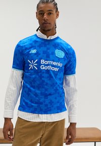 Blaues Sporttrikot mit einem pixelartigen Muster und weißen Logos. Darüber getragen wird ein weißes Nadelstreifenhemd, kombiniert mit braunen Cordhosen.