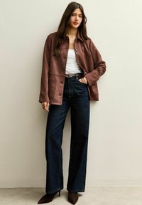 Chaqueta de ante marrón con cierre de botones y dos bolsillos delanteros, combinada con jeans de mezclilla oscuros de tiro alto y zapatos de tacón marrón puntiagudos.