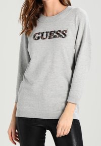 Šedá mikina oversized střihu s nápisem "GUESS" v dekorativních aplikacích na prsou. Hranaté manžety a spodní lem.