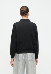 Μαύρο φούτερ με γιακά, μακριά μανίκια και ριμπ τελειώματα στα μανίκια. Η πίσω πλευρά έχει λεία υφή με ελάχιστες ραφές.