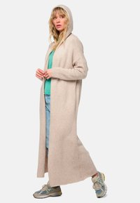 Long cardigan beige avec capuche, à l'avant ouvert et aux manches longues, fabriqué dans un matériau doux. Porté sur un haut vert et associé à un denim clair.