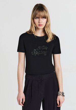 Giovane donna con capelli biondi indossa una maglietta nera a maniche corte con il design di perline "Hello Spring" e pantaloni neri larghi, in posa contro uno sfondo bianco.