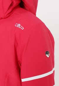 Épaule de veste rouge avec broderie du logo blanc "CMP", badge en métal et bande réfléchissante horizontale blanche sur la manche.