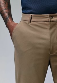 Beige Hosen mit glatter Textur, ausgestattet mit Seitentaschen und einem Knopfverschluss. Neben einem dunkelblauen gemusterten Oberteil präsentiert.