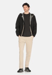 Giacca nera con cappuccio e fodera interna grigia, con chiusura a zip, indossata sopra una maglietta scura e pantaloni della tuta beige, abbinati a sneakers nere.