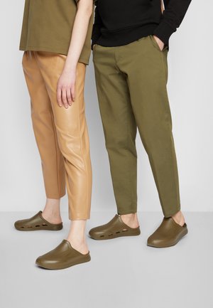 Duas pessoas estão lado a lado, vestindo calças de pele sintética bege, calças de algodão verde e sapatos slip-on escuros de clogs com orifícios de ventilação.