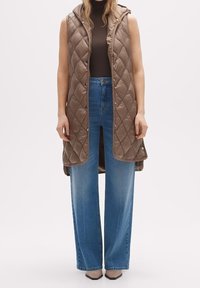 Brun vadderad väst med huva, framtida tryckknappar, bärs över en mörk polo och ljusblå vida jeans. Slät och glänsande textur.