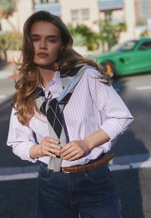 Młoda kobieta o długich włosach, ubrana w pasiastą koszulę i sweter zarzucony na ramiona, stojąca na ulicy z zielonym samochodem sportowym w tle.