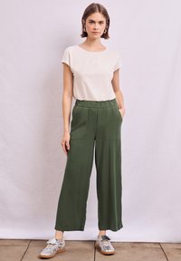 Witte short-sleeve top gecombineerd met hoge taille, wijde groene broek met zijzakken. Model draagt zilveren sneakers.