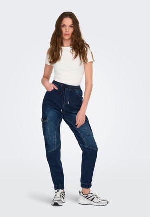 Woman standing with one hand in pocket, wearing white short-sleeve top, dark denim cargo joggers, and white athletic shoes.