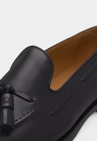 Fekete bőrből készült férfi mokaszin bojt részlettel és világosbarna belső béléssel, közelről, ferdén felülről nézve.