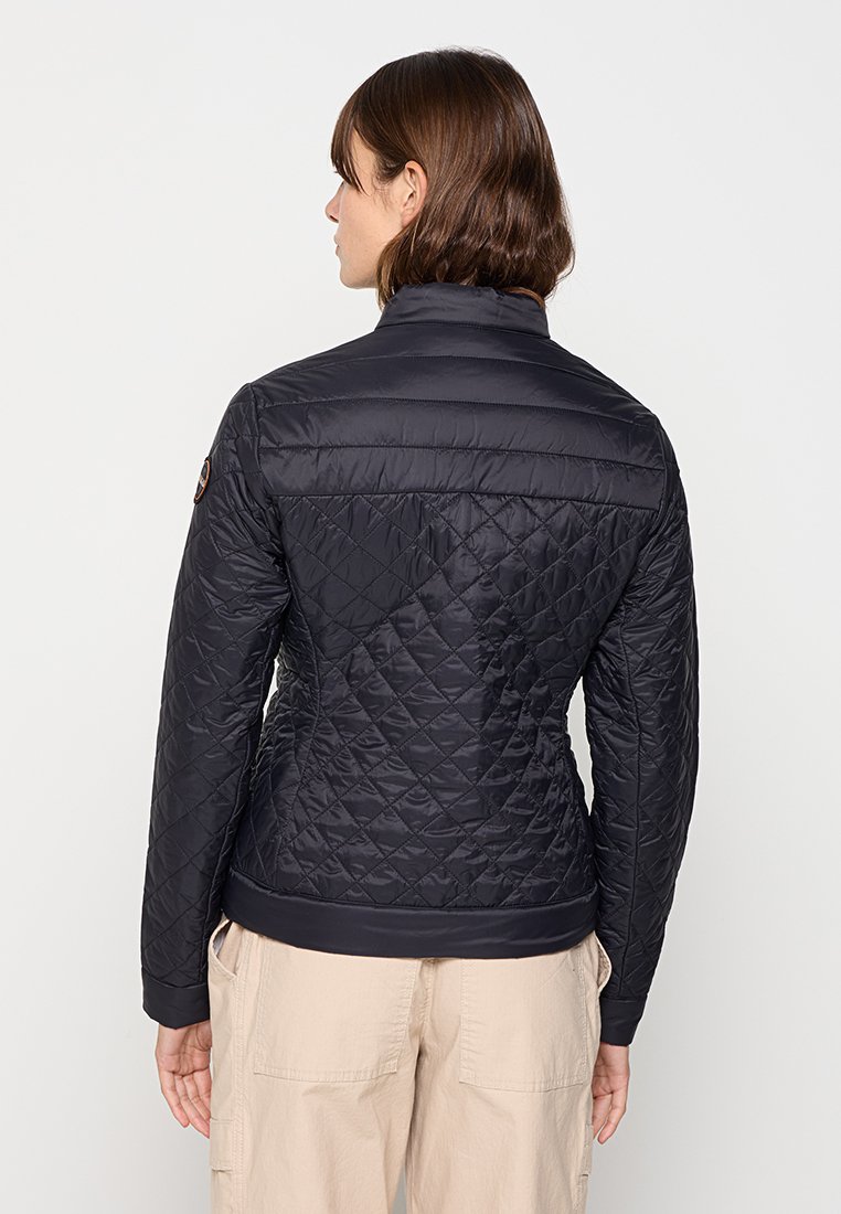 Frau mit kurzen braunen Haaren, die eine figurbetonte schwarze gesteppte Jacke und beigefarbene Hose trägt, von hinten vor einem schlichten Hintergrund gezeigt.