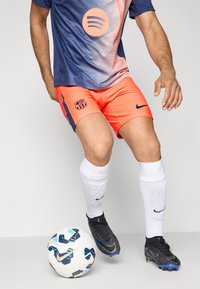 Nogometni igrač u plavoj i koraljnoj opremi s bijelim čarapama kontrolira bijelu i tamnoplavu Nike nogometnu loptu na sivoj pozadini.