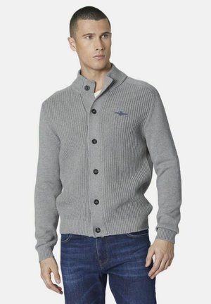 Cardigan grigio a costine con colletto a scialle, chiusura a bottoni e bottoni scuri. Presenta un piccolo logo sul petto. Indossato con jeans blu.