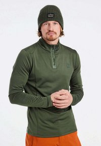 Grünes Langarm-Zip-Up-Oberteil mit hohem Kragen, versehen mit einem kleinen Logo und strukturierten Ärmeln, kombiniert mit einer olivfarbenen Strickmütze.