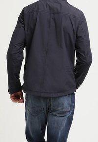 Marineblå, lättviktsjacka med en slät yta, dragkedjefickor och ståkrage. Bakifrån visar den rena linjer och minimal detaljer.