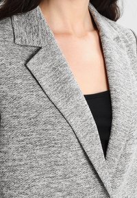 Grau gestrickter Blazer mit Reverskragen, strukturierten Finish und lockerer Silhouette, über einem schwarzen Oberteil getragen.