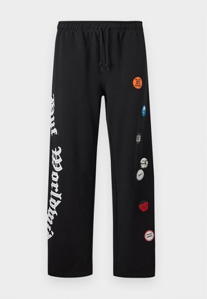 Man draagt zwarte joggingbroek met witte grafische tekst op het linkerbeen en diverse kleine patches, gecombineerd met zwart-witte sneakers.