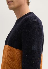 Breit gebreide trui met een donkerblauwe bovenkant en een gestructureerd oranje ondergedeelte, geaccentueerd met multicolore vezels en een logo-opdruk op de mouw.
