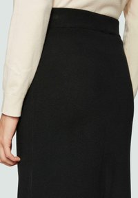 Geribbelde zwarte rok met een hoge tailleband, en een gladde textuur. Gepaard met een lichtgekleurde lange mouw top, hand rustend op de rok.