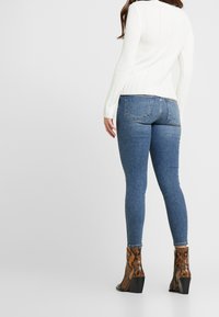 Kvinna som bär en vit tröja, blå smala jeans och bruna mönstrade ankelstövlar står mot en enkel bakgrund och vänder sig bort.