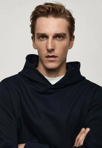 Modelka oblečená v tmavomodrej mikine s jemnou textúrou, dlhými rukávmi a predným vreckom, na neutrálne pozadí.