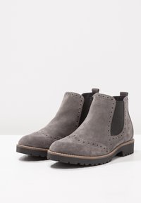 Grå mockaskor med ankellängd, elastiska sidopaneler, brogue-detaljer längs tån och en låg svart gummisula. Rundad tådesign.