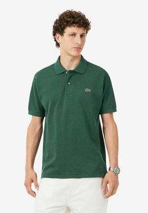 Joven con una camiseta tipo polo de color verde oscuro con un pequeño logo en el pecho y pantalones blancos, posando contra un fondo liso.