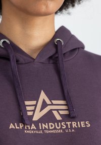 Lila kapucnis pulóver pamutból, fém szemekkel, bézs grafikai kialakítással és "ALPHA INDUSTRIES" logós szöveggel az elején.