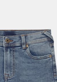 Modré džínové kalhoty s vysokým pasem, s měděným knoflíkem, přední kapsou a viditelným prošitím. Látka má vybledlou texturu.