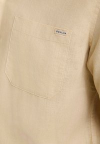 Chemise en lin beige avec une poche appliquée portant une étiquette indiquant "PULLIN". Le tissu a une surface légèrement texturée.
