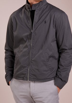 Mann trägt eine dunkelgraue Jacke mit Reißverschluss, hohem Kragen und Seitentaschen, kombiniert mit hellgrauer Hose, Hände in den Taschen.