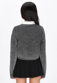 Gestrickter grauer Pullover mit einem verkürzten Design, geripptem Saum und langen Ärmeln, kombiniert mit einem schwarzen, plissierten Rock. Weißer Kragen ist sichtbar.